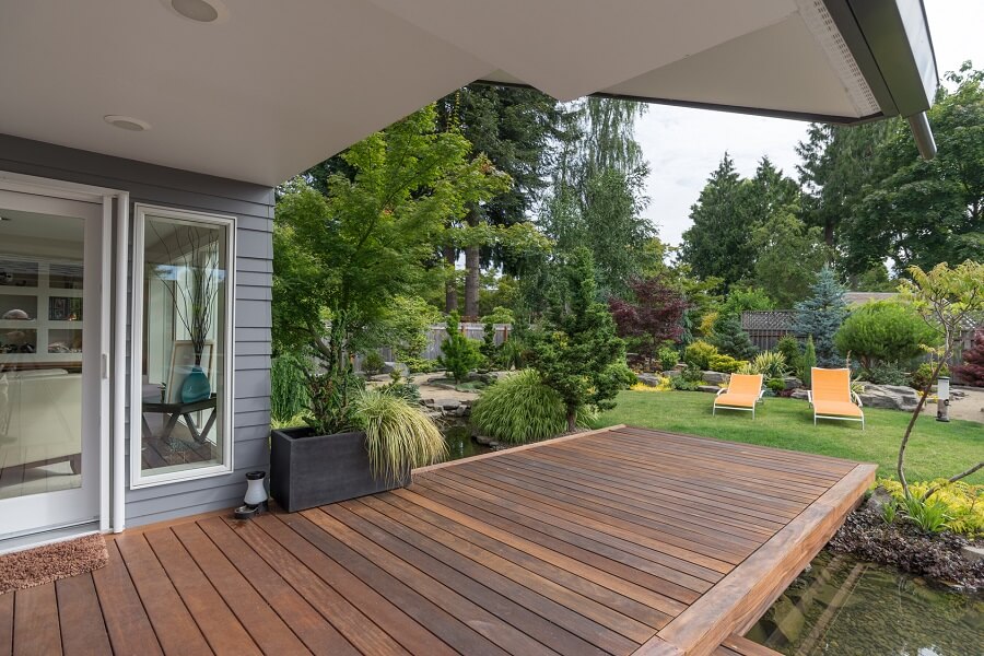 A perspective view of a contemporary Pacific Northwest home with a deck bridging a pond that leads to a pair of modern yellow loungers in a landscaped yard.
